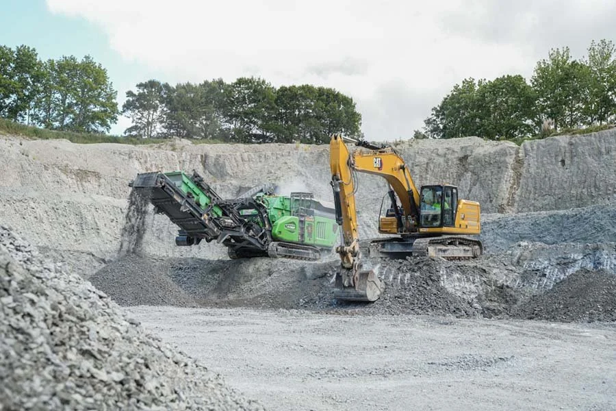 A yellow excavator working alongside a green rock crusher in a quarry, with trees in the background.