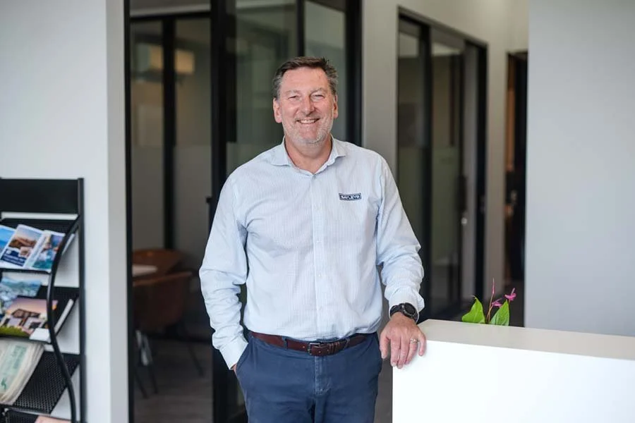 A smiling man in a white button-up shirt and blue pants standing in an office, leaning on a white counter with a pink flowering plant on it.