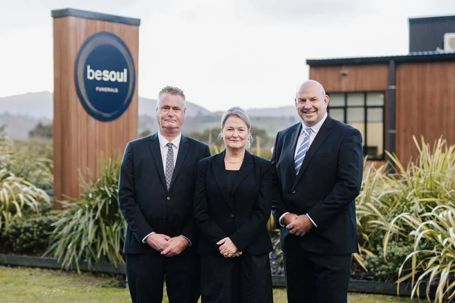 Three professionally dressed people, two men and one woman, standing outside in front of a building with a sign that reads 'besoul funerals.' They are smiling and the background shows a modern building and green landscaping.