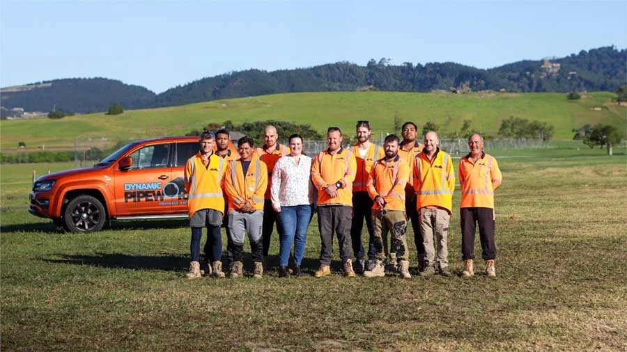 Group of ten people standing outdoors in front of an orange vehicle with 'Dynamic Pipe' logo on it, with green hills and trees in the background.