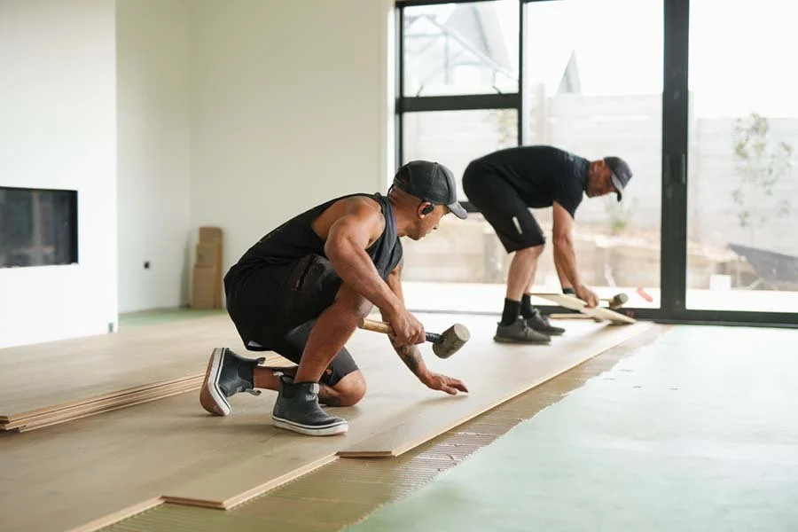 Two men installing hardwood flooring in a bright room with large glass windows.