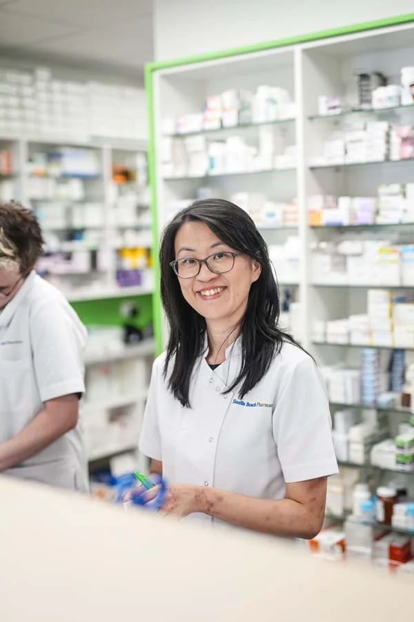 Pharmacist smiling in a pharmacy with shelves of medicines in the background.