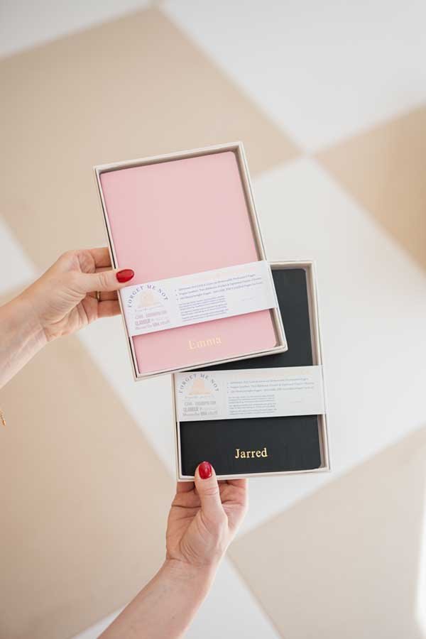 Two boxed notebooks, one pink with the name Emma and one black with the name Jarred, being held by a person with red nail polish in a room with a beige and white checkered floor
