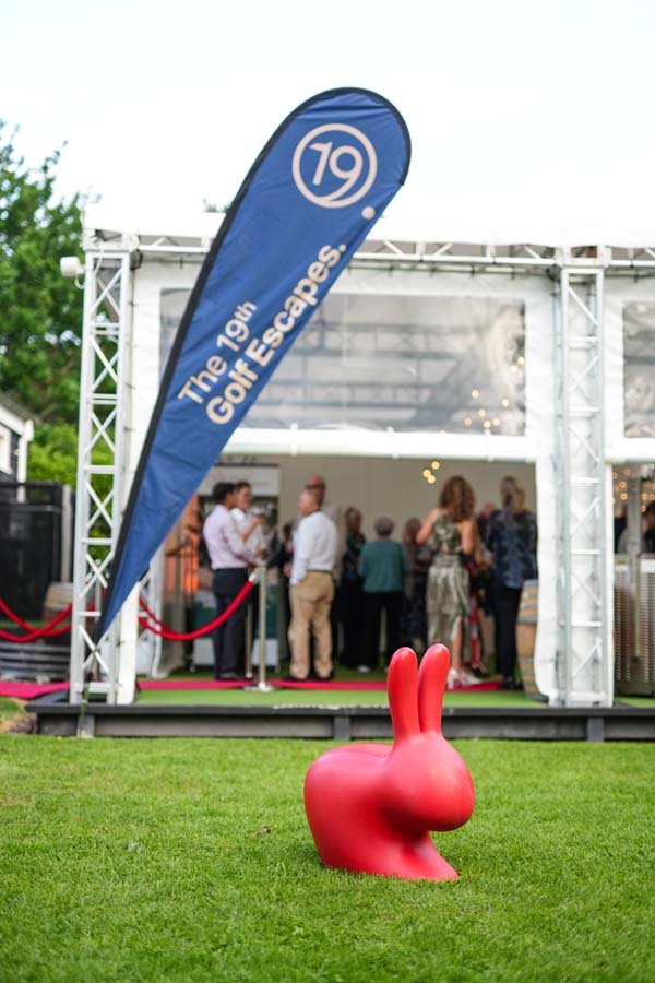A red Bunny sculpture on green grass in front of a white event tent with people gathered inside, and a blue flag reading 'The 19th Golf Escapes'