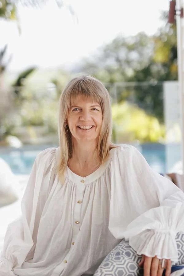 A woman with blonde hair smiling, wearing a white blouse, sitting outdoors with a pool and greenery in the background.