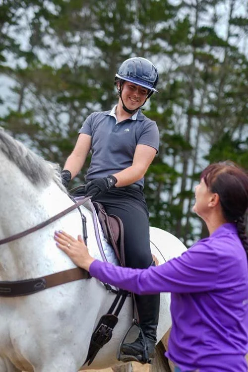 A woman wearing a riding helmet and gloves is sitting on a white horse, smiling. Another woman dressed in a purple jacket is standing beside the horse, reaching up to touch its side and talking to the rider. Trees are visible in the background.