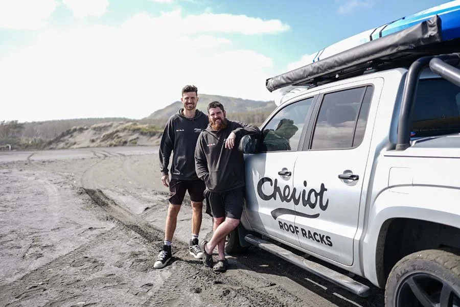 Two men standing next to a white truck with an outdoor landscape in the background. One man is leaning on the truck, smiling, while the other is standing nearby. The truck has the text 'Chariot Roof Racks' on the side.