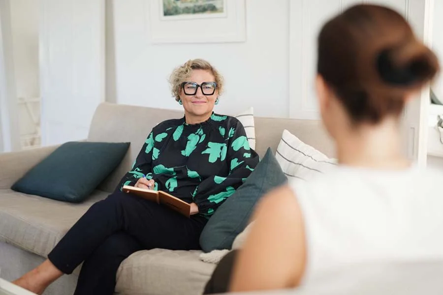A woman with curly gray hair, large glasses, and earrings sitting on a beige couch with dark and striped pillows, talking to another woman with her back to the camera in a bright, cozy room.