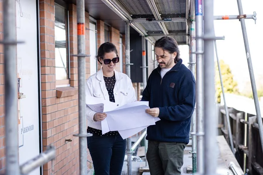 A woman and a man review blueprints on a construction site with scaffolding outside a brick building.