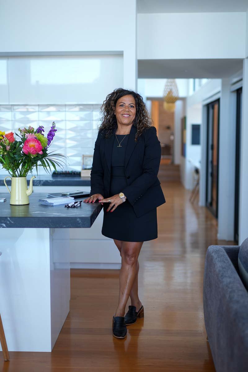 A woman with curly hair wearing a black blazer and dress, standing in a modern kitchen near a countertop with a yellow pitcher holding colorful flowers, smiling at the camera.
