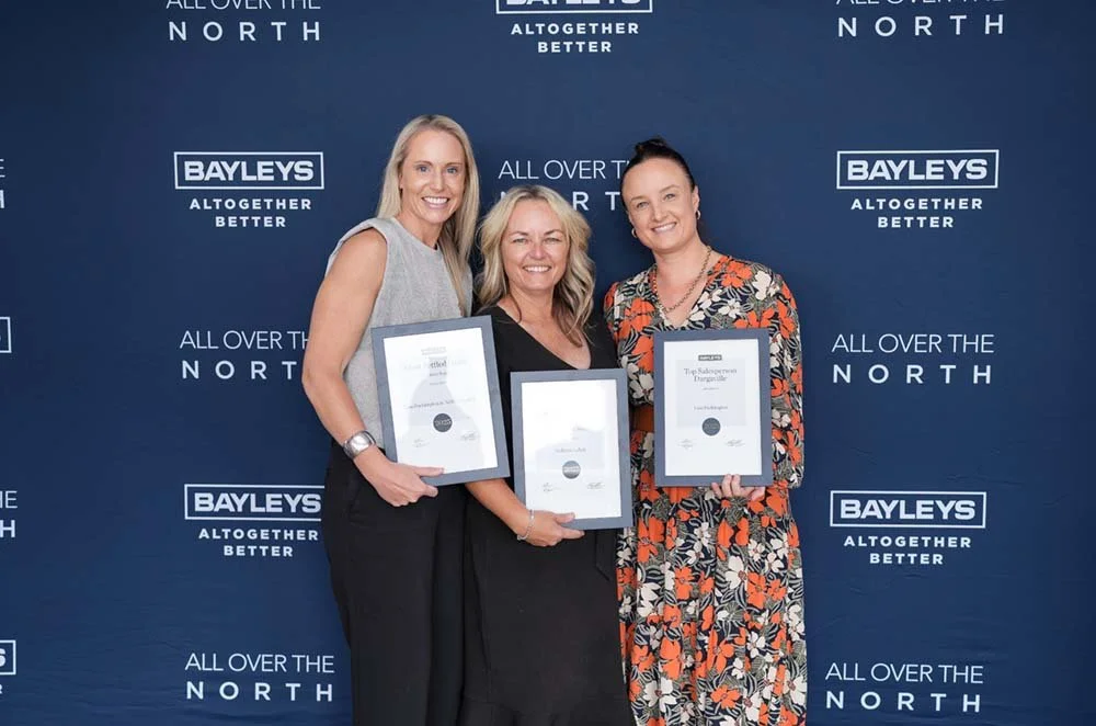 Three women standing in front of a blue backdrop with Bayleys and All Over The North logos, holding framed certificates and smiling.