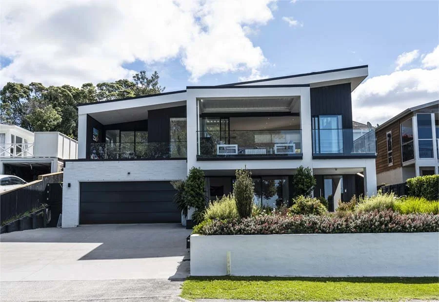 Modern two-story house with black and white exterior, large glass windows, a balcony with seating, and a landscaped front yard with bushes and flowers.