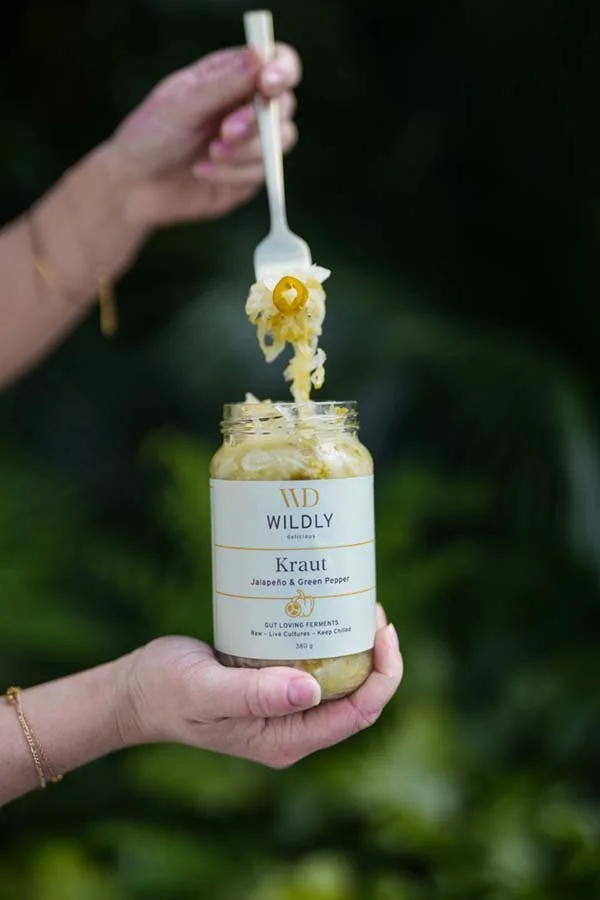 Person holding a jar of fermented sauerkraut with a fork scooping out some kraut, labeled 'WILDLY Kraut Jalapeño & Green Pepper' against a blurred green outdoor background.