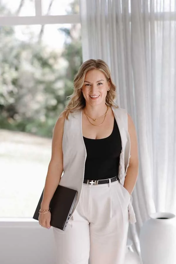 A smiling woman with blonde hair, wearing a black top, white pants, and a light blazer, holding a closed laptop, standing indoors near a window with curtains, with a blurred outdoor garden visible outside.