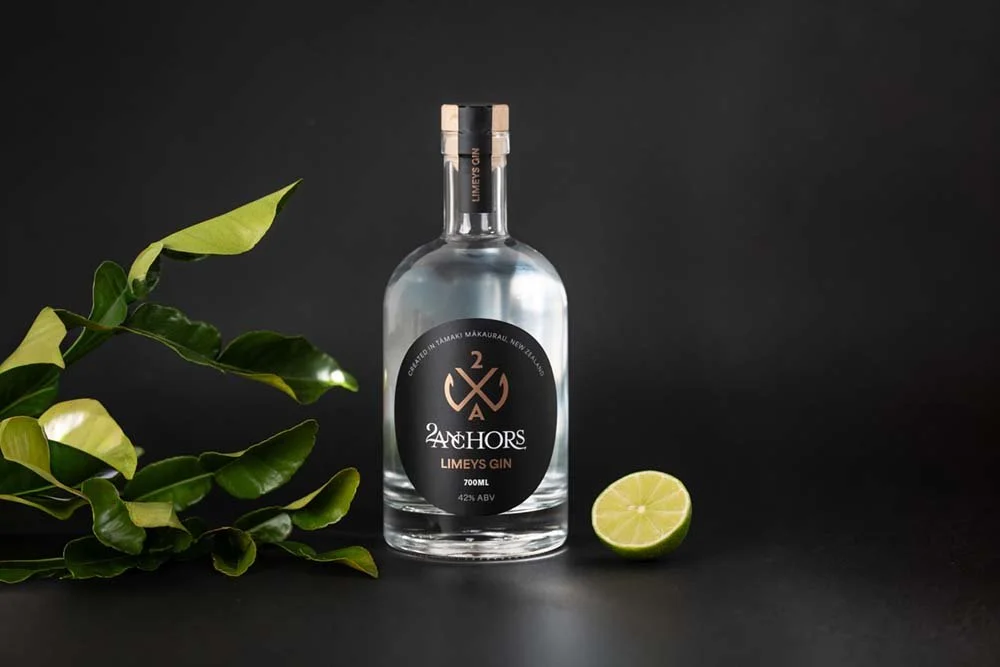 A bottle of Anchors Limeys Gin with a lime half and leafy plant on black background.