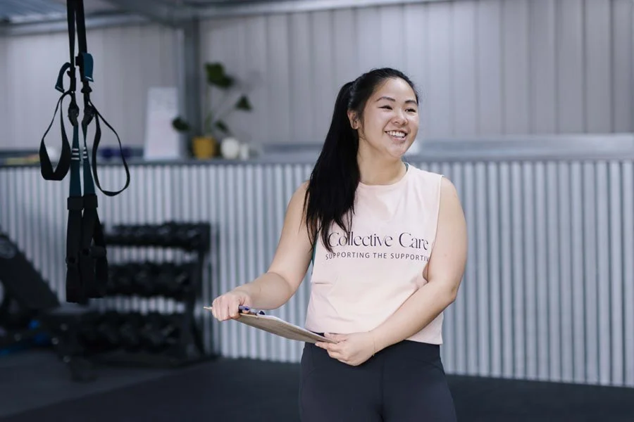 A smiling woman holding a clipboard in a gym with suspension training equipment hanging from the ceiling.