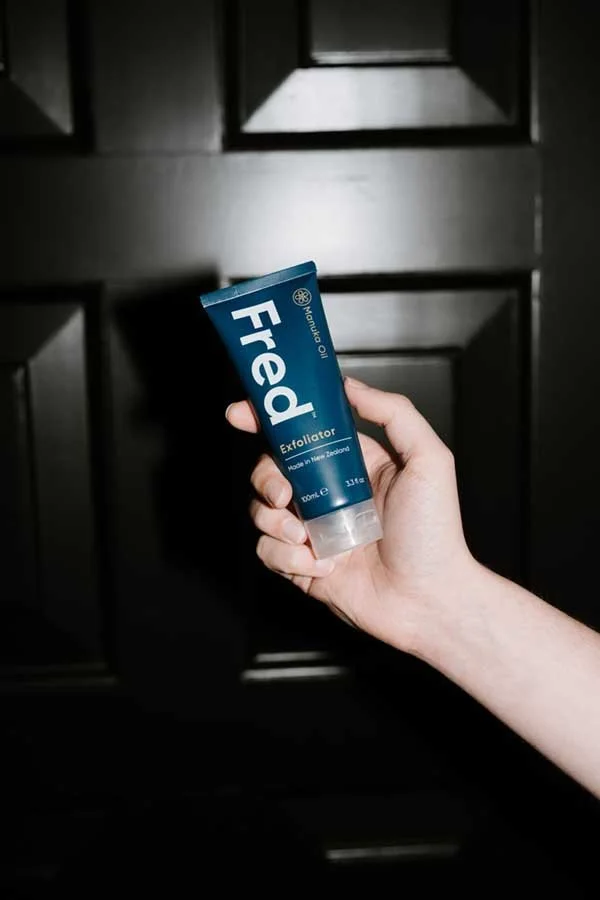 Hand holding a tube of exfoliating face scrub labeled "Fred" in front of a dark wooden door.