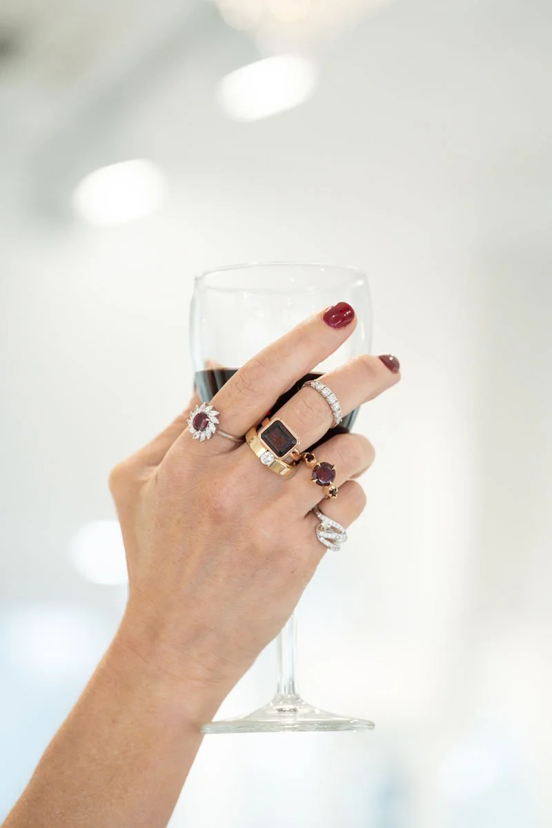 Hand holding a glass of red wine, adorned with multiple rings including a large square ring, a diamond ring, and other decorative rings.
