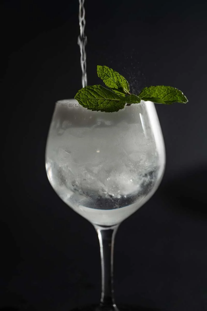 A wine glass filled with a clear beverage and ice, garnished with a sprig of fresh mint leaves, with water being poured into the drink.