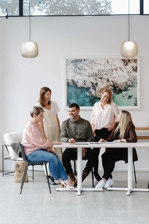 Five people gathered around a white table in a modern office, with two hanging pendant lights above and a large aerial photograph on the wall behind them. They are smiling and engaging in conversation.