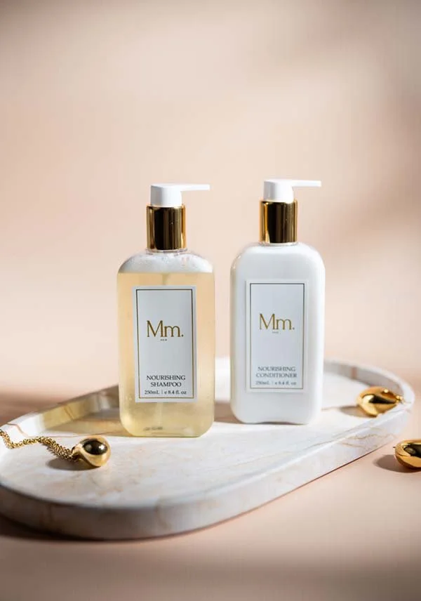 Two bottles of hair care products, a nourishing shampoo and conditioner, placed on a white marble tray with gold accents and jewelry, against a light beige background.