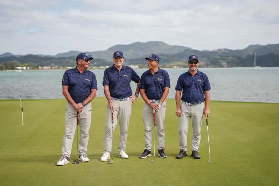 Four men in matching navy blue golf shirts and light-colored pants standing on a golf course near a body of water, holding golf clubs, with hills in the background.