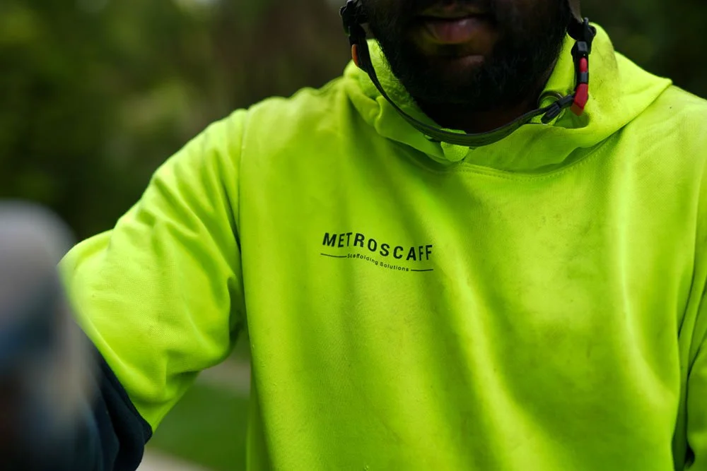 Close-up of a person wearing a neon yellow METROSCFF cycling shirt and black helmet, outdoors with green foliage in the background.
