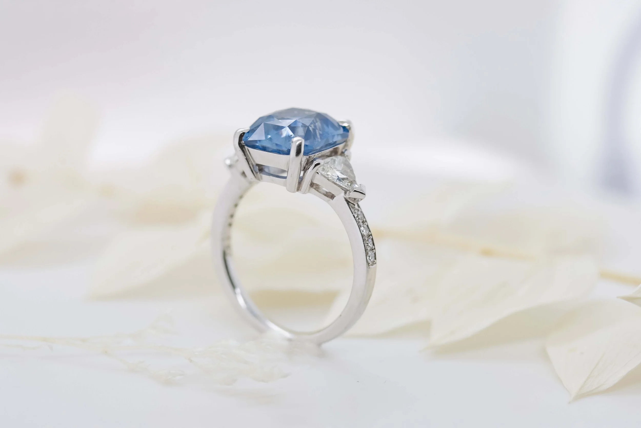 A silver ring with a large blue gemstone and smaller clear stones, placed on white petals.