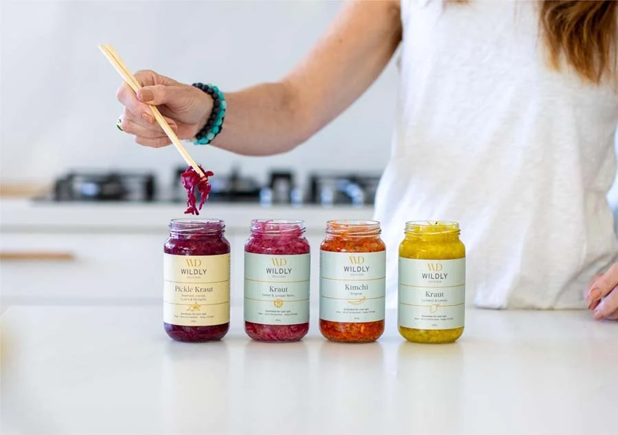 A person scooping purple kimchi with chopsticks from a jar, with three other jars of different fermented vegetables on a white countertop in a kitchen.