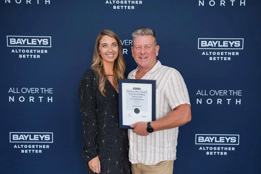 A man and woman smiling and posing together, with the man holding a framed award certificate. They are standing in front of a blue backdrop with the Bayleys logo and the phrase 'All over the North'.