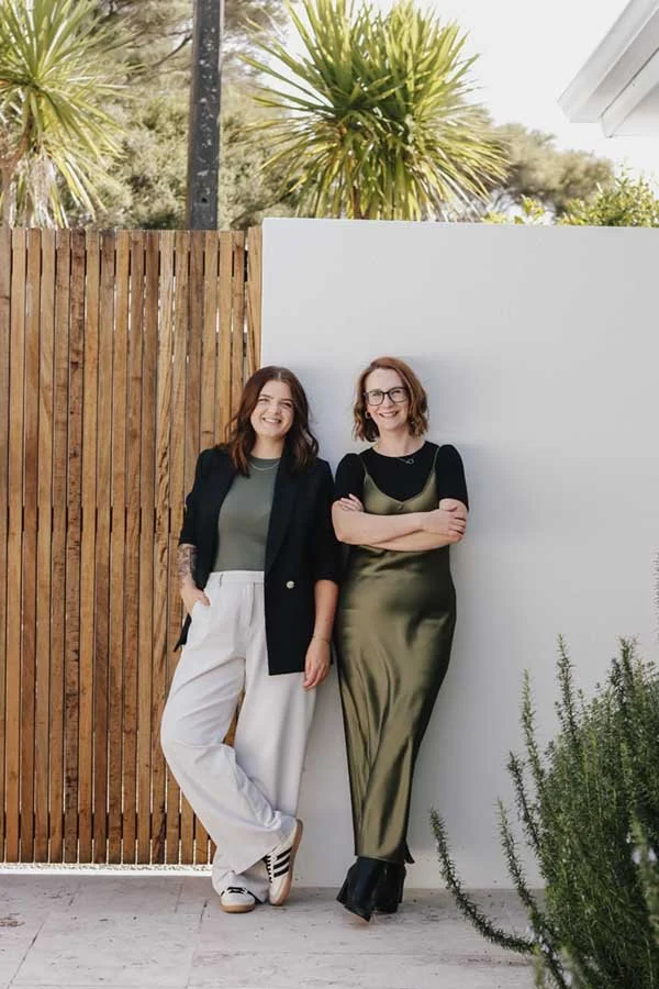 Two women standing outdoors, smiling, with a wooden fence and white wall behind them, and palm trees in the background.
