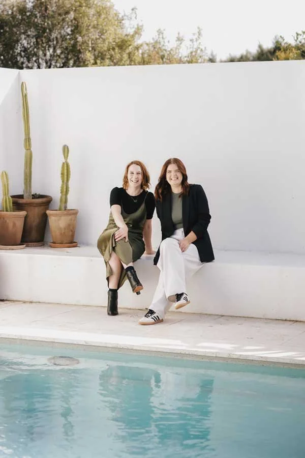Two women sitting by a pool on a white ledge, smiling, with tall potted cacti and a white wall behind them, outdoors during daytime.