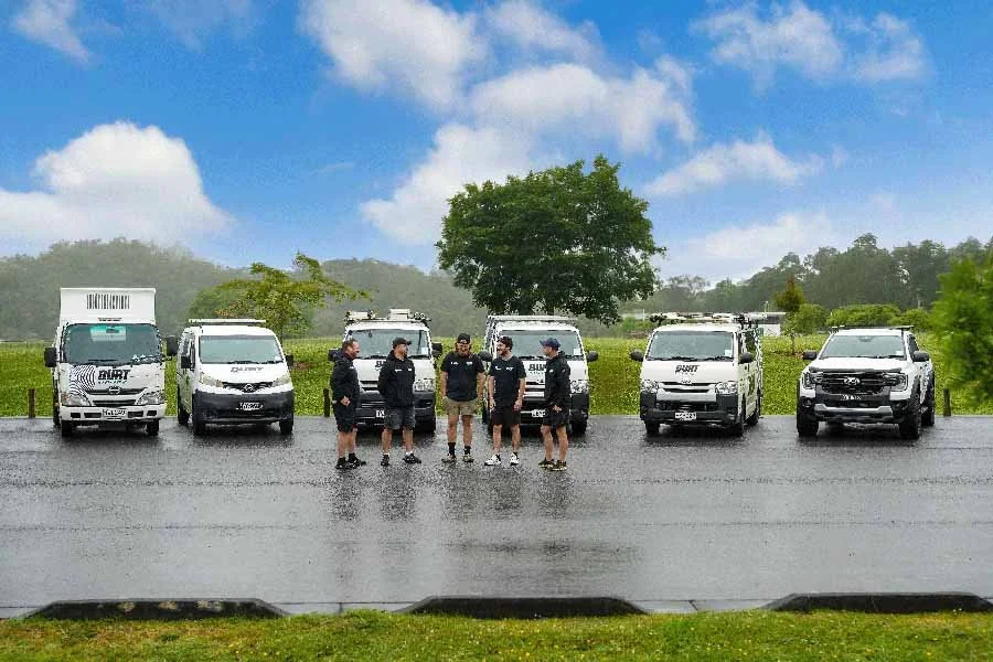Group of six police officers standing and talking in a parking lot with six police vehicles parked behind them on a rainy day.