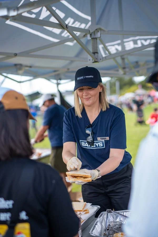 A woman wearing a navy blue Batley's shirt, black cap, and gloves is serving a sandwich at an outdoor event under a canopy. There are people and tents in the background.