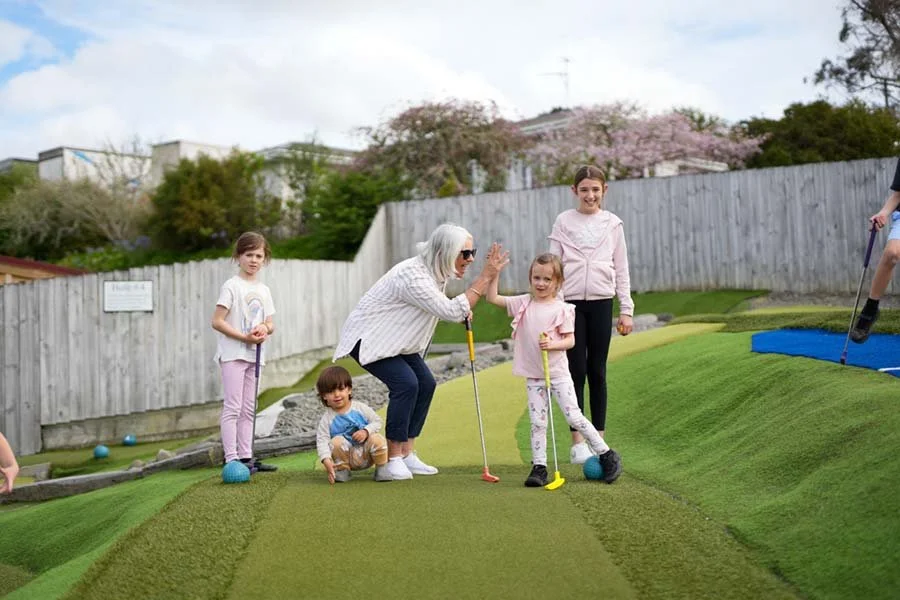 A group of children and an elderly woman playing mini-golf outdoors on a sunny day.
