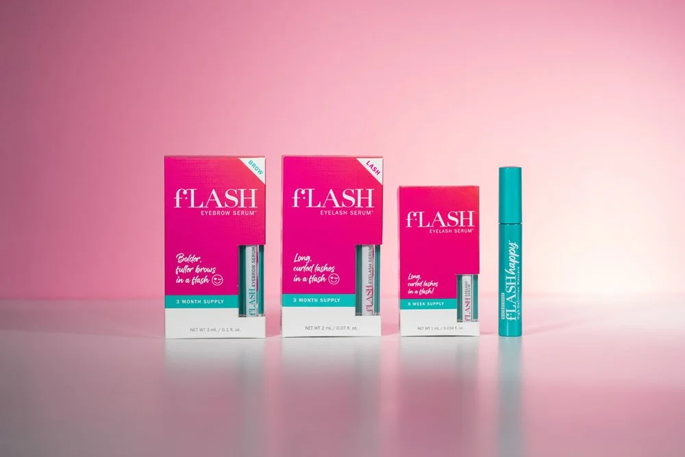 Three boxes and one cylindrical container of FLASH eyelash serum on a pink background. The boxes are pink with teal accents, and the serum tube is teal with white text.
