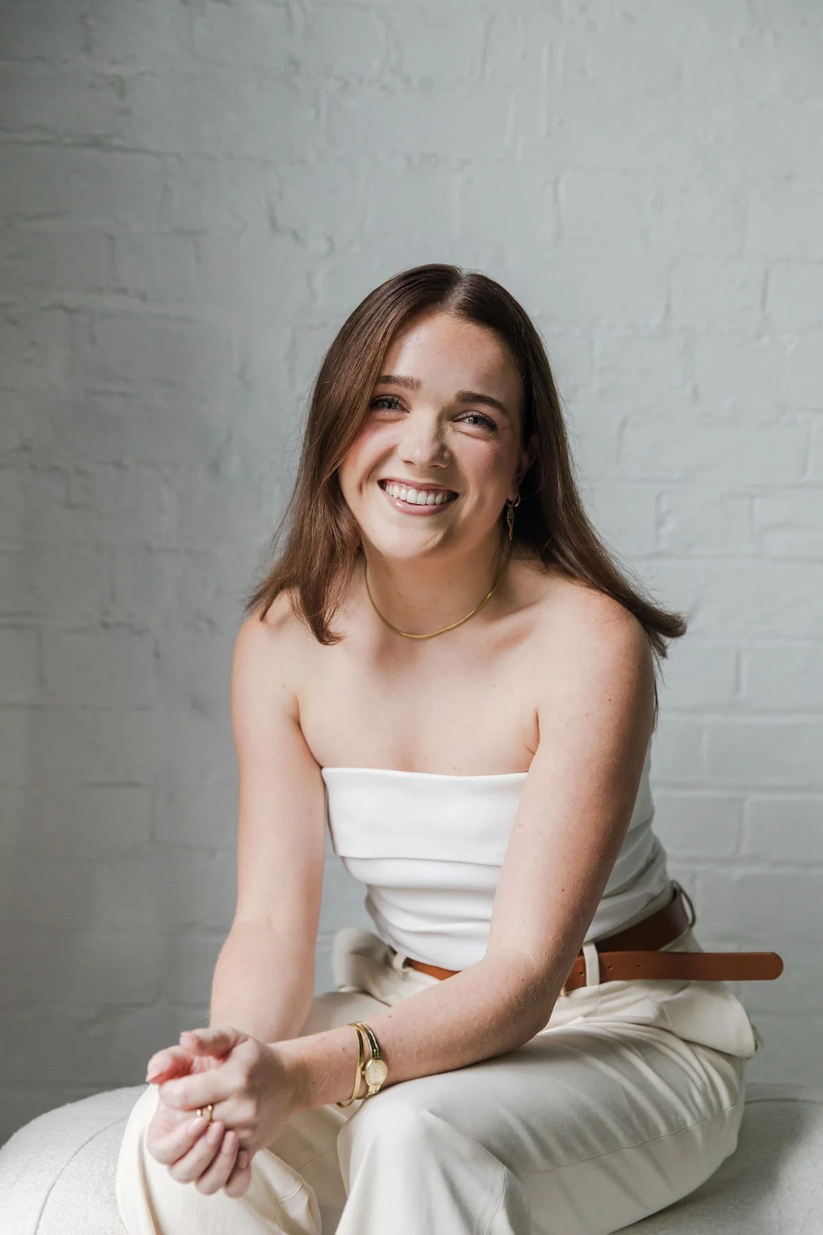 A young woman with shoulder-length brown hair, smiling, sitting against a light gray brick wall, wearing a white strapless top, cream-colored pants, a gold necklace, a gold wristwatch, and a bracelet.