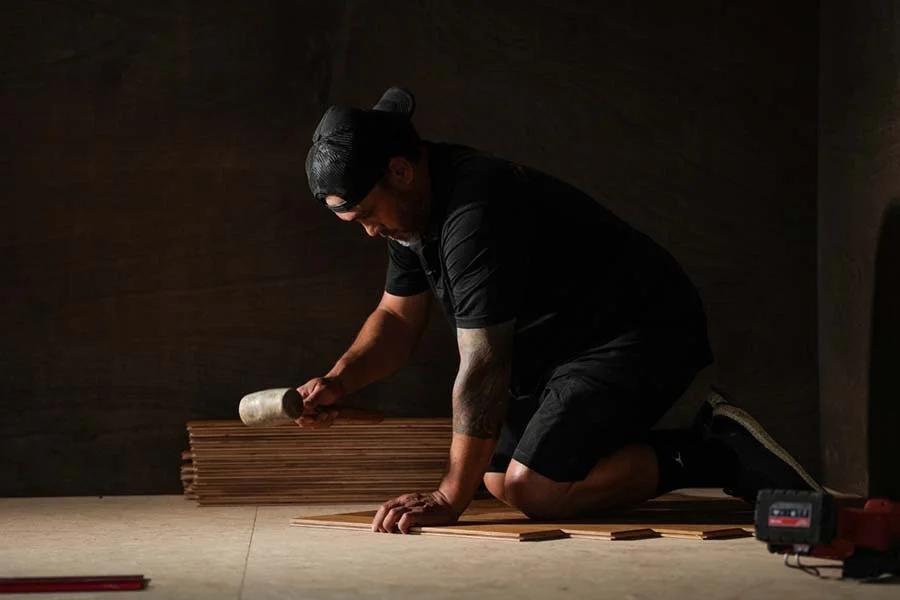 A man wearing a black cap, black shirt, and shorts is kneeling on the floor, using a mallet to tap wooden planks laid out in a row for flooring installation.