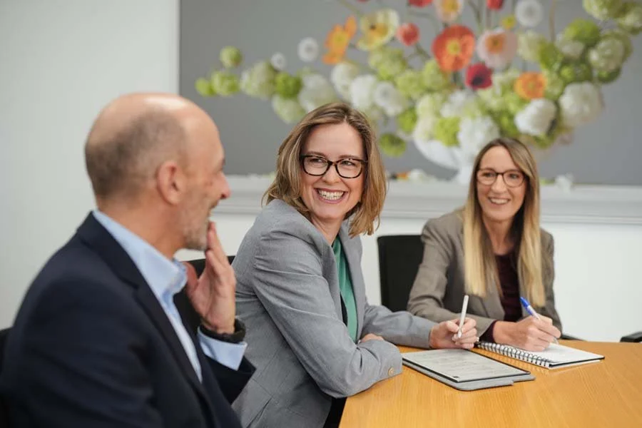 Three business professionals, two women and one man, engaged in a meeting in a conference room. The woman in the middle is smiling and wearing glasses, the woman on the right is taking notes, and the man on the left is listening. A flower painting de