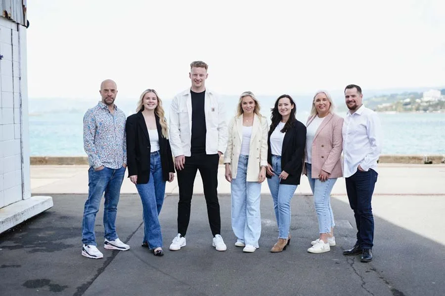 Seven diverse adults standing outdoors by a body of water on a clear day, smiling at the camera.