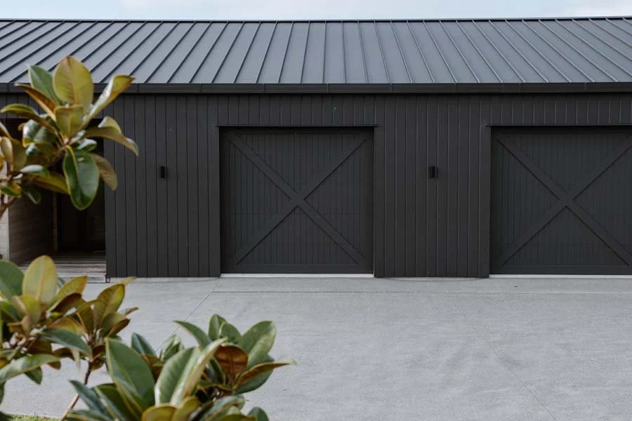 Black double garage doors on a building with a metal roof, with green and brown leaves in the foreground.