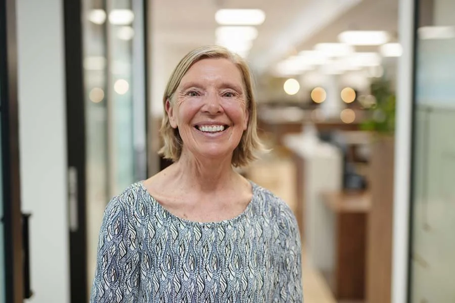 A smiling middle-aged woman with blonde hair, wearing a patterned blue and white blouse, standing indoors in a well-lit, modern office or commercial space.