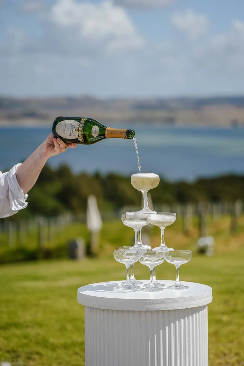 Person pouring champagne into a glass on top of a pyramid of champagne glasses outdoors with a lake and cloudy sky in the background.