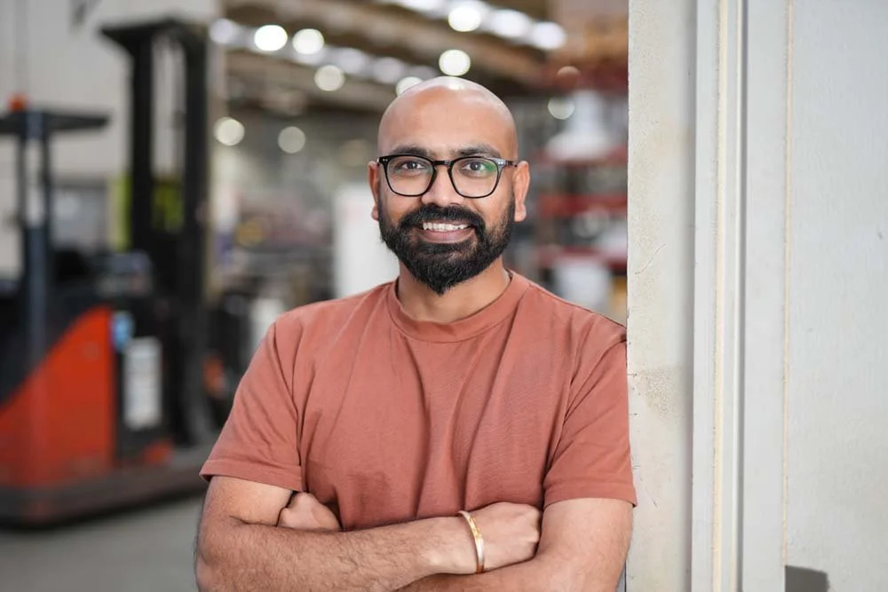 A man with glasses and a beard smiling, standing with arms crossed in a workshop or industrial space.