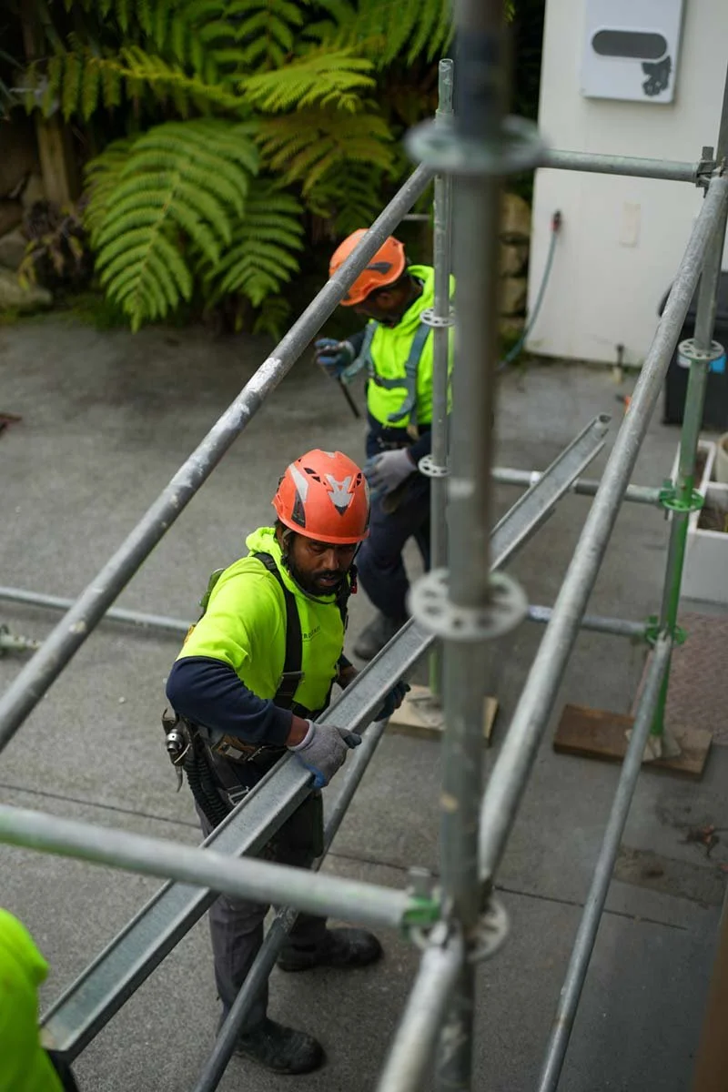 Two construction workers wearing orange helmets and bright yellow safety vests working on scaffolding outdoors. One worker is looking at the camera while holding onto the scaffold, and the other is in the background, looking at their phone. Large gre