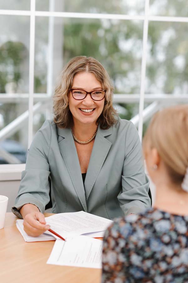 A professional woman with glasses and a blazer smiling during an interview or consultation with a woman with blonde hair. They are seated at a table with papers and a pen.
