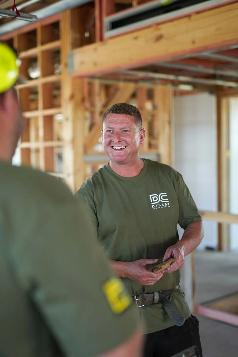 Two construction workers inside a wooden building frame, smiling and talking. One worker is partially visible in the foreground, and the other is fully visible, holding some tools.