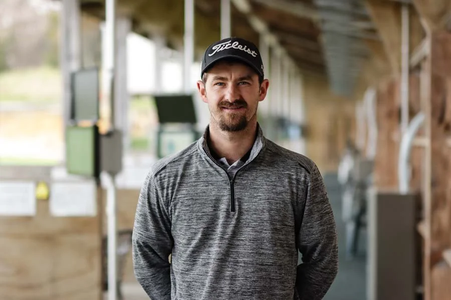A man in a gray quarter-zip sweater and a black Titleist golf cap standing indoors with a wooden interior, possibly a golf clubhouse or facility.