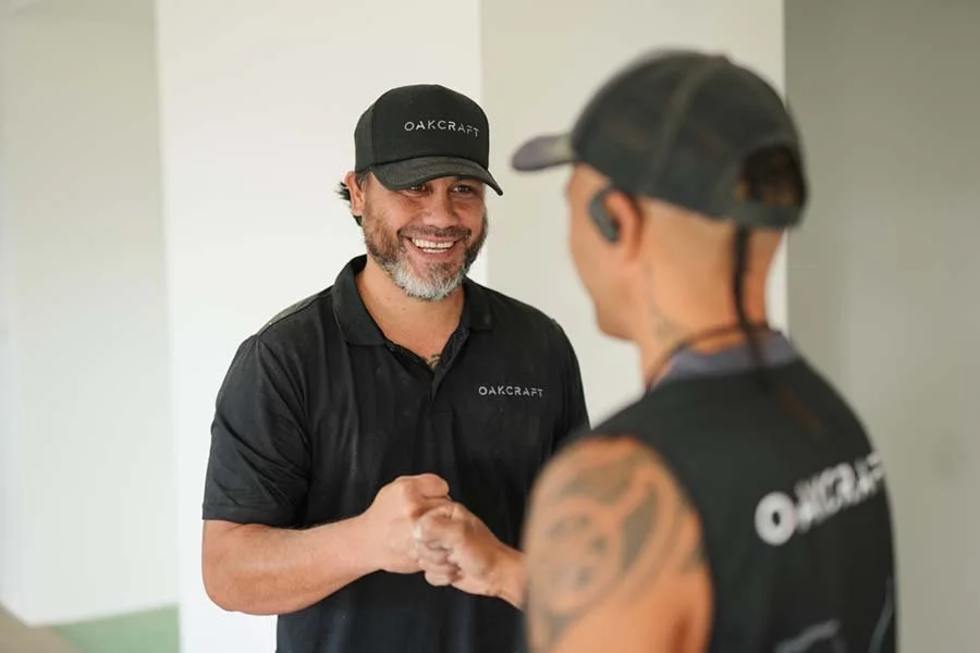 A man with a beard wearing a black Oakcraft shirt and cap shaking hands with a young man in a black Oakcraft tank top and cap.