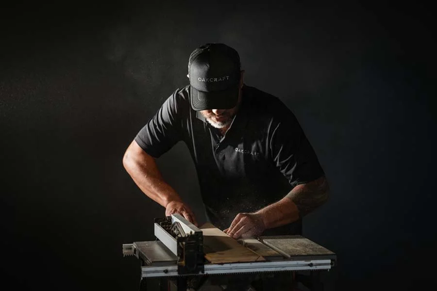 A man working with a saw in a dark room, wearing a black hat and shirt, concentrating on a woodworking project.